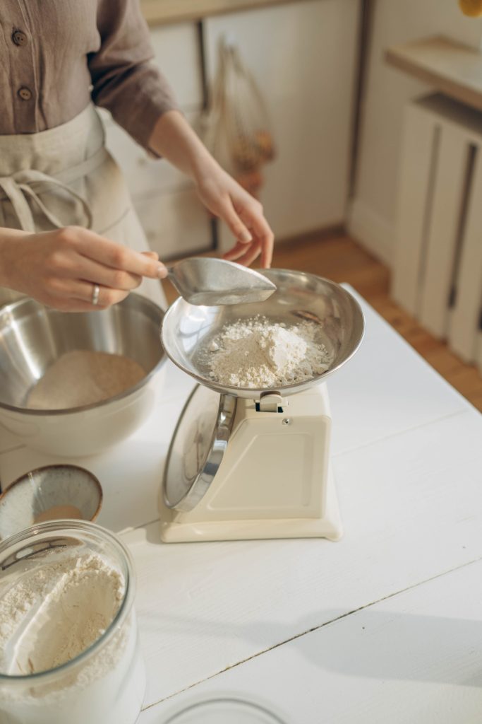 A person measuring flour for baking using a vintage weighing scale on a white table.