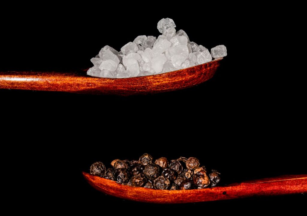 Artistic arrangement of salt and pepper on wooden spoons, contrasting textures and colors on a black background.
